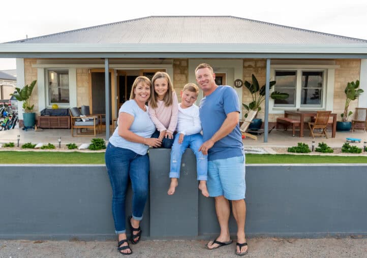 outdoors portrait of a happy family of four smiling in front of new dream home or vacation rental house. mom, dad, and children boy and girl, embracing and having fun together enjoying holiday villa.