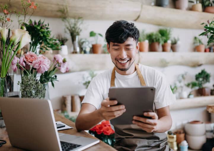 asian male florist, owner of small business flower shop, using digital tablet while working on laptop against flowers and plants. checking stocks, taking customer orders, selling products online. daily routine of running a small business with technology