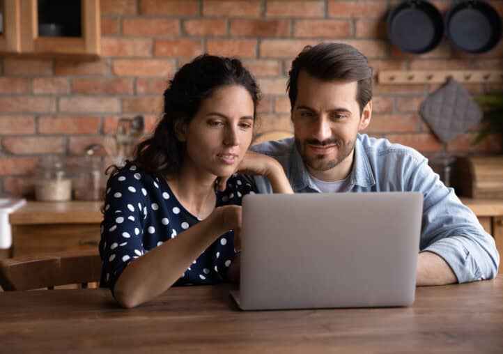 happy millennial couple using laptop in kitchen, watching show together