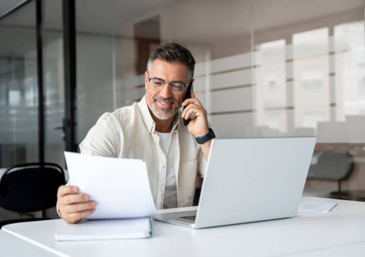 middle aged latin businessman having call on smartphone with business partners or clients. smiling mature hispanic man sitting at table talking by mobile cellphone, using laptop, paper documents