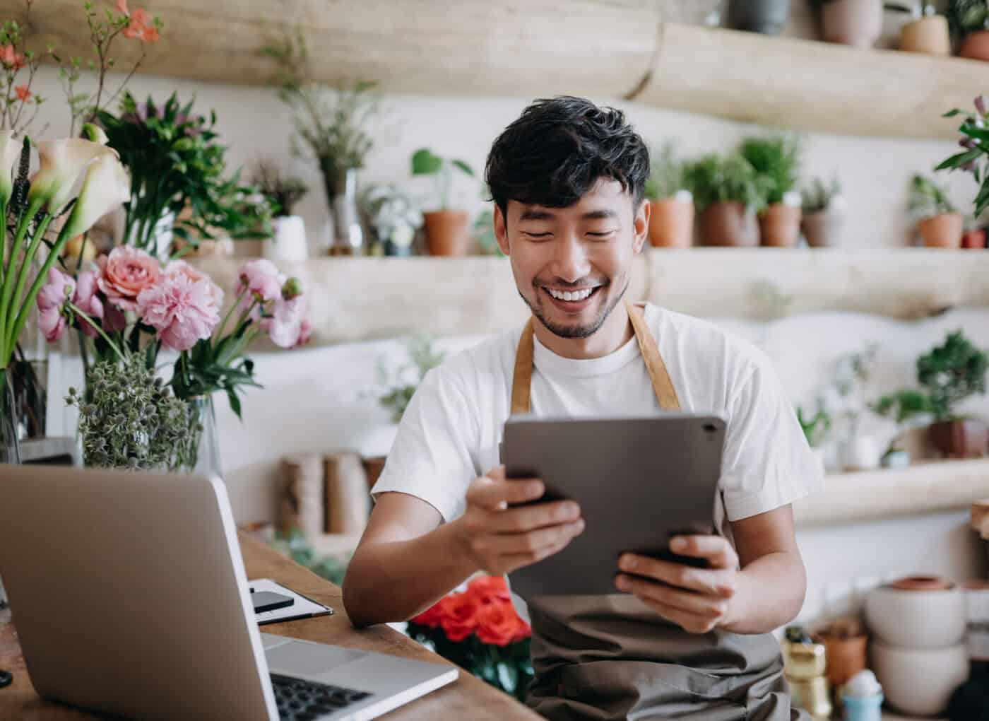 asian male florist, owner of small business flower shop, using digital tablet while working on laptop against flowers and plants. checking stocks, taking customer orders, selling products online. daily routine of running a small business with technology