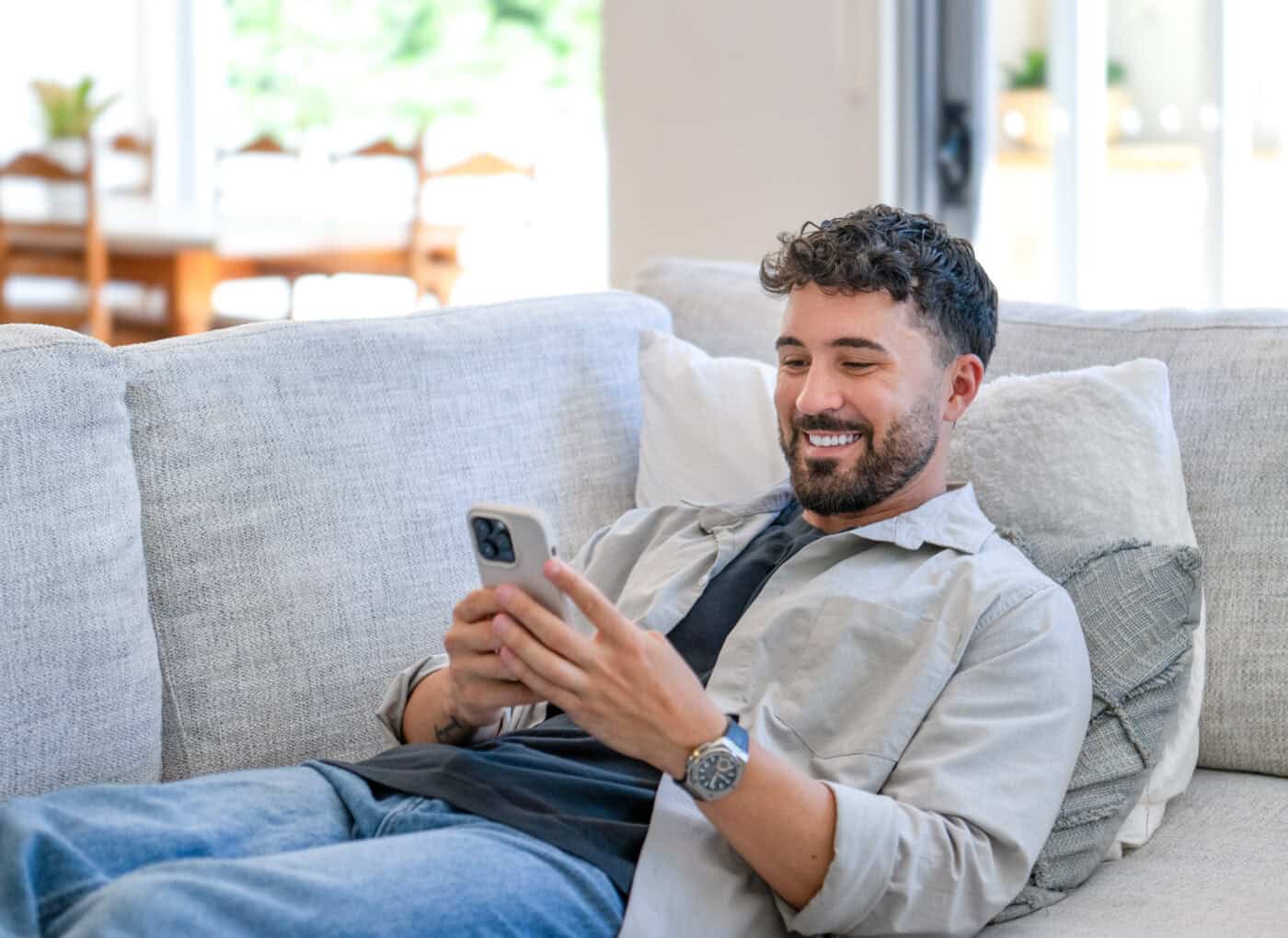 happy young man relaxing sitting and reclining on the sofa at home. he is using a mobile phone.