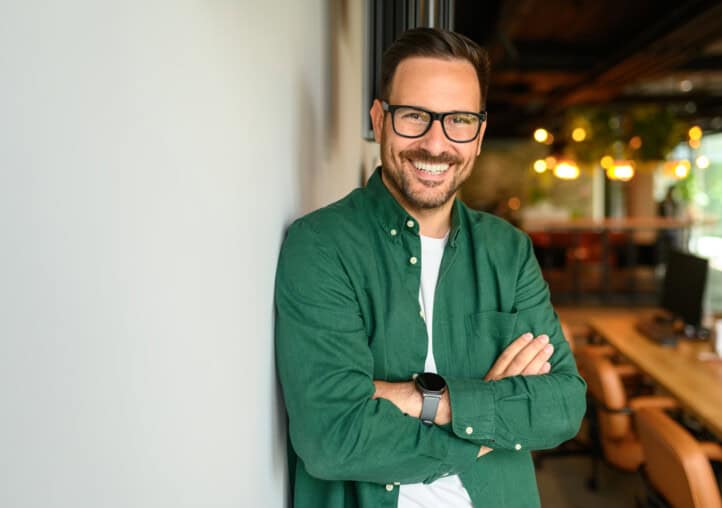 portrait of confident businessman in glasses and with arms crossed leaning on white wall in office